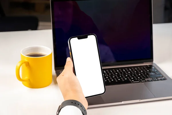 Video call mock up, close up woman hand holding vertical smartphone video call mock up.  Office worker female drinking coffee, take a break from using laptop and looking white blank screen of phone.