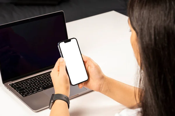 Holding smartphone, woman sitting on the desk and holding smartphone. Horizontal modern mobile phone mock up. Caucasian female using laptop and attending online meeting on her personal cellphone.