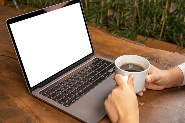 Modern laptop, woman drinking coffee and looking modern laptop. White blank screen mock up. Sitting wooden table, attending online meeting. Watching movie. E-learning concept idea.