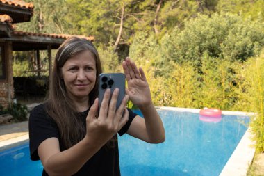 Video çağrısı, mutlu sarışın kadın portresi. Tatilde akıllı telefonla görüntülü konuşma yapmak. Tatilde çalışmak, yüzme havuzunun önünde durmak, online başvuru kullanmak, el sallamak..