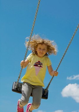 Çocuk yüksekten sallanıyor. Arka planda mavi gökyüzü var. Portre biçimi.