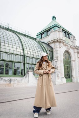 Beyzbol şapkalı genç bir kadın çatıda dururken trençkotun kemerini ayarlıyor. 