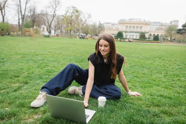 Gülümseyen Genç Kadın Çimenlerde Otururken Dizüstü Bilgisayar Kullanıyor — Stok fotoğraf
