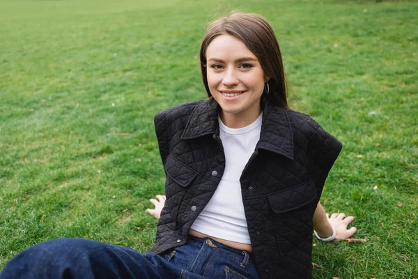Çimenlerde Oturan Kolsuz Ceketli Gülümseyen Genç Kadın — Stok fotoğraf