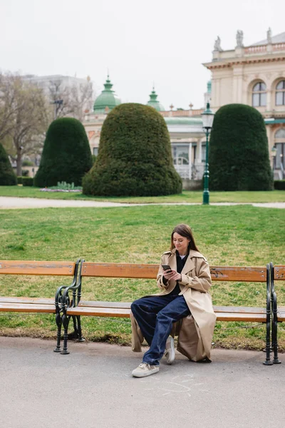 young woman in stylish trench coat smiling and using smartphone