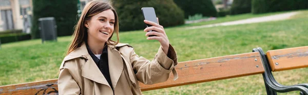 young woman in stylish trench coat taking selfie outside, banner