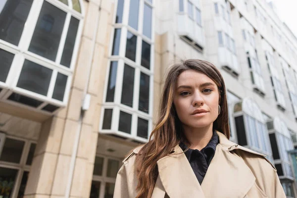 low angle view of young woman in stylish trench coat looking at camera near building