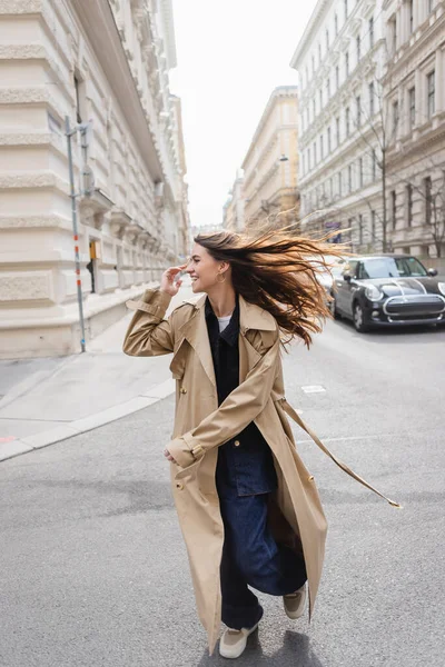 happy young woman in trench coat looking away while walking on street in windy day