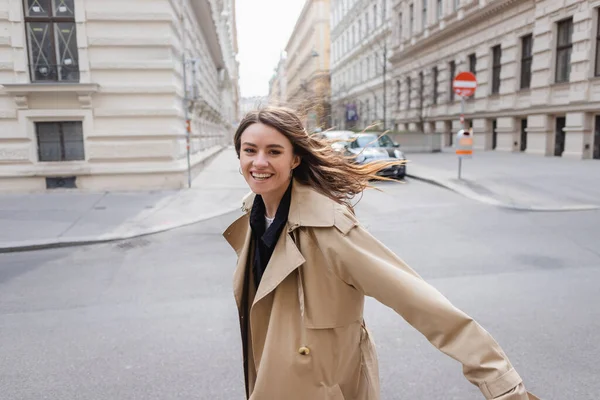 cheerful young woman in trench coat walking on street in windy day