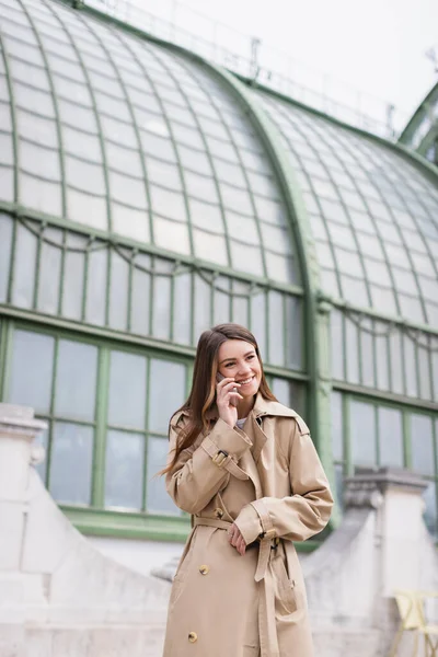 happy young woman in trench coat talking on smartphone near austrian building