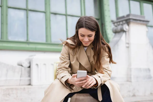 stock image joyful woman in stylish trench coat using mobile phone near european building 