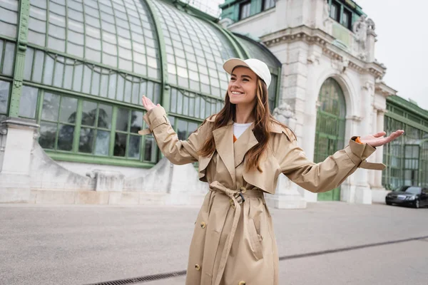 happy young woman in trench coat and baseball cap standing with outstretched hands on rooftop of european building