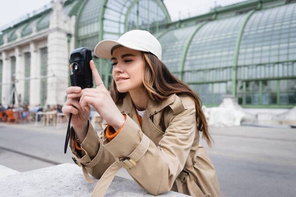 young woman in beige trench coat and baseball cap taking photo on vintage camera 