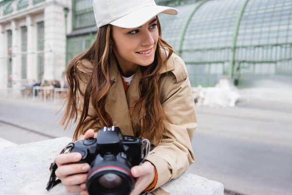 happy photographer in trench coat and baseball cap holding digital camera 