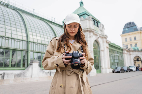 photographer in trench coat and baseball cap holding digital camera in european city