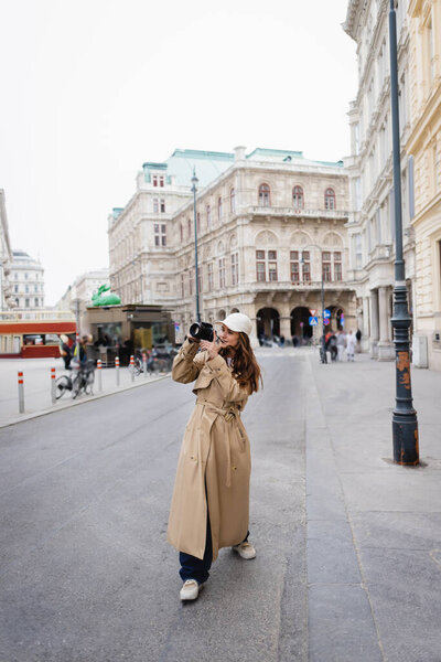 full length of young woman in stylish trench coat and baseball cap taking photo on digital camera in european city