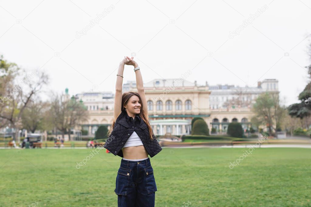 young cheerful woman in sleeveless jacket stretching in green park 
