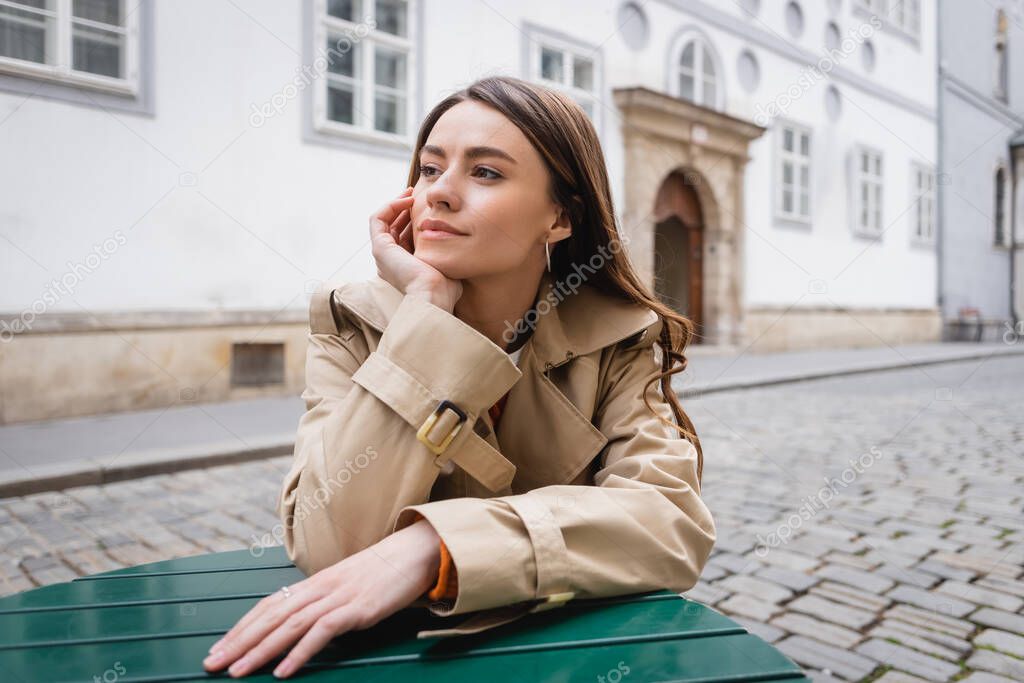 dreamy young woman in trendy trench coat sitting in summer terrace 