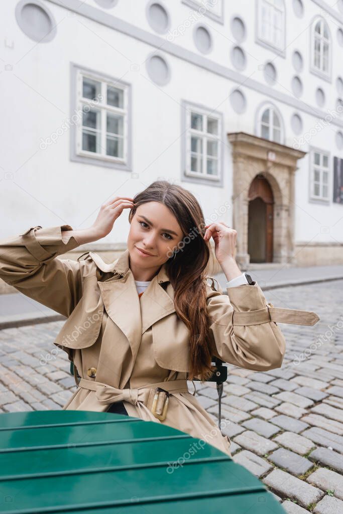 young woman in trendy trench coat adjusting hair and sitting in summer terrace 