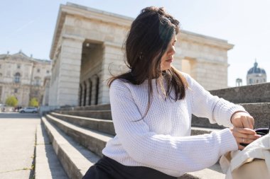 Esmer kadın beton merdivenlerde otururken sırt çantasını açıyor. 