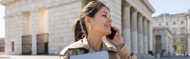 Trençkotlu neşeli kadın dizüstü bilgisayarını tutuyor ve dışarıda akıllı telefondan konuşuyor.
