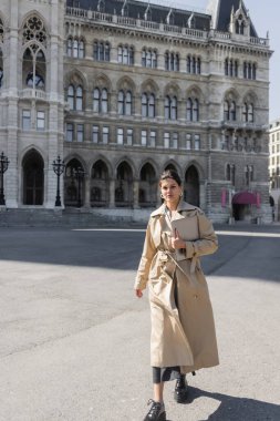 stylish freelancer in beige trench coat walking with laptop on street of vienna  clipart