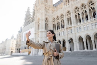 Bej renkli trençkotlu serbest çalışan Viyana Belediye Binası yakınlarında dizüstü bilgisayar tutarken selfie çekiyor. 
