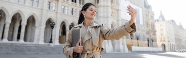 Bej renkli trençkotlu serbest çalışan Viyana belediye binasının yanında dizüstü bilgisayar tutarken selfie çekiyor.