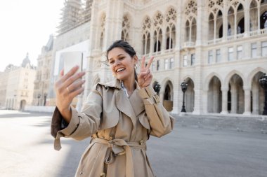 Bej renkli trençkotlu mutlu kadın barış işareti gösteriyor. Viyana belediye binasının yakınında selfie çekiyor.