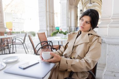 Trençkotlu kadın kafenin terasında akıllı telefon ve dizüstü bilgisayarın yanında müzik dinliyor.