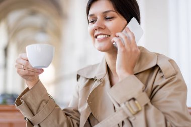 Yazlık terasta elinde fincanla akıllı telefondan konuşan neşeli bir kadın. 