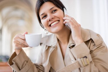 Yazlık terasta elinde bardak tutan ve akıllı telefondan konuşan gülümseyen bir kadın. 