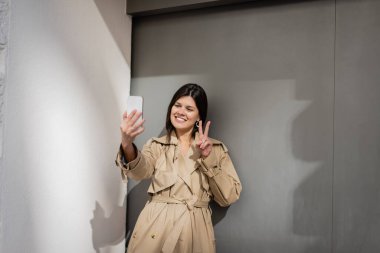 Trençkotlu mutlu kadın selfie çekiyor ve duvarın yanında barış işareti gösteriyor. 