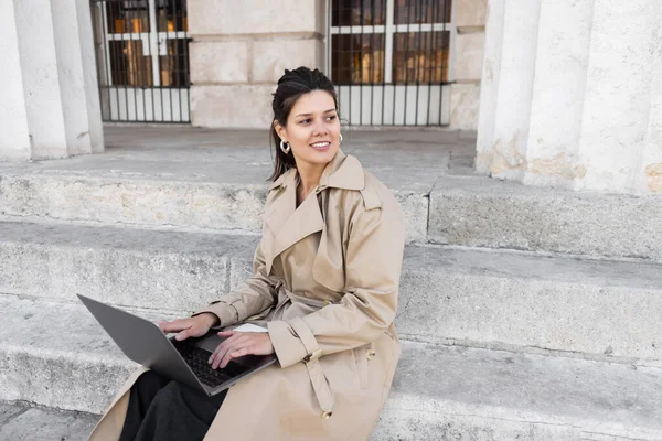 pleased woman in trench coat sitting on concrete stairs and working remotely on laptop