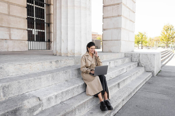веселый фрилансер в плаще сидит на бетонной лестнице, разговаривает на смартфоне и удаленно работает на ноутбуке в Вене