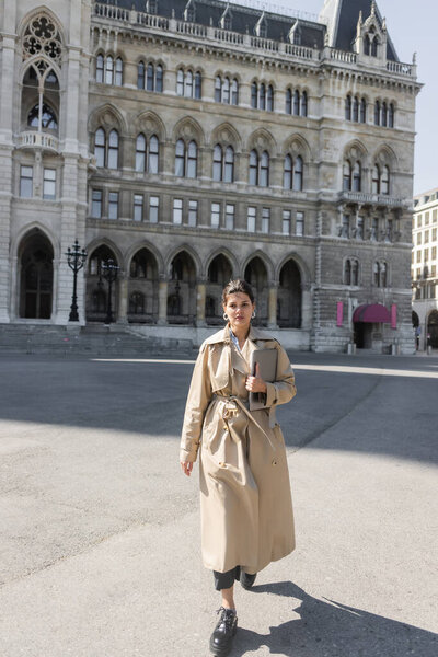 фрилансер в модном бежевом плаще прогуливается с ноутбуком по улице Виенны 