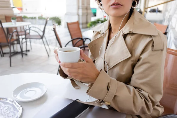 トレンチコートに身を包んだ女性が音楽を聴きながらカフェテラスでスマートフォンやノートパソコンの近くでカップを持ち — ストック写真