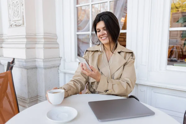 cheerful woman in trench coat listening music and holding cup while taking photo on smartphone in cafe terrace