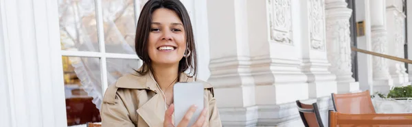 smiling woman in trench coat listening music in earphones and holding smartphone in cafe terrace, banner