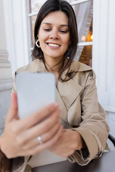 Trençkotlu Gülümseyen Kadın Kafenin Terasında Bulanık Akıllı Telefon Tutuyor — Stok fotoğraf