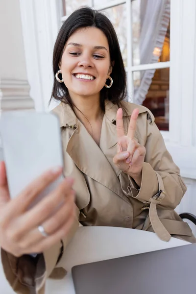 トレンチコート姿の明るい女が平和サインを出しカフェテラスのぼやけたスマートフォンで自撮り — ストック写真