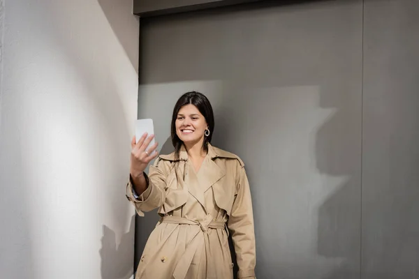 穿着风衣的快乐女人在墙边的智能手机上自私自利 — 图库照片