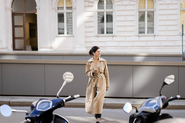 smiling woman in wired earphones holding mobile phone and listening music while wlaking on street in vienna