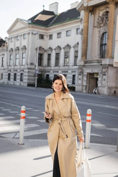 Usmívající Žena Stylovém Kabátě Drží Tašku Smartphone Ulici Vídni — Stock fotografie