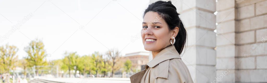 brunette and smiling woman in beige trench coat looking at camera, banner