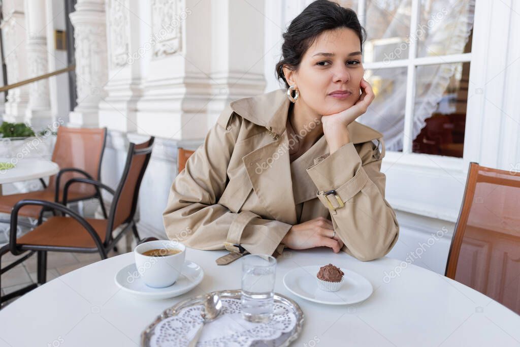 brunette woman in trench coat sitting near cupcake and cup of coffee in cafe terrace