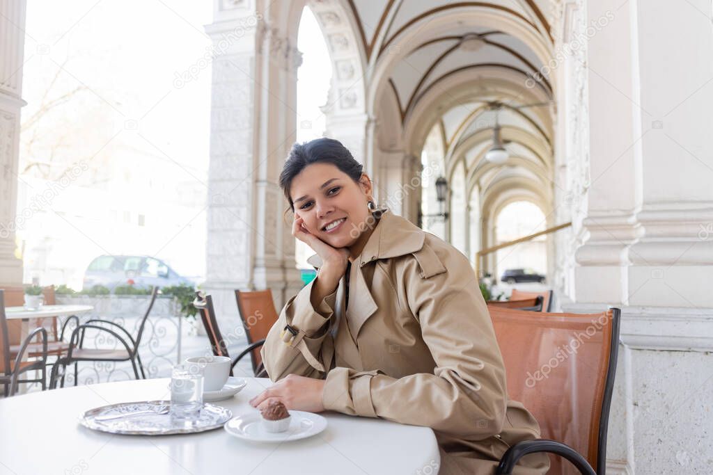 cheerful woman in trench coat sitting near cupcake and cup of coffee in cafe terrace