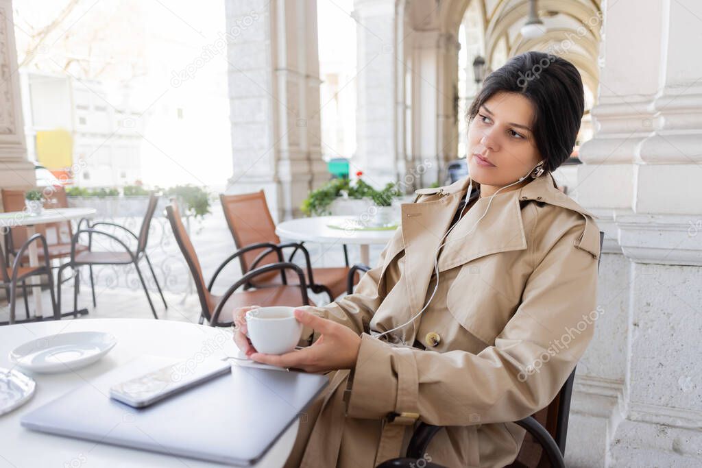 woman in trench coat listening music near smartphone and laptop in cafe terrace