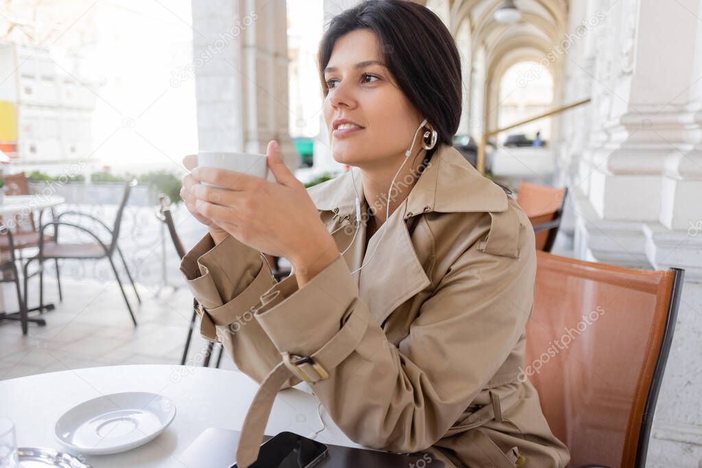 happy woman in trench coat listening music and holding cup near smartphone and laptop in cafe terrace