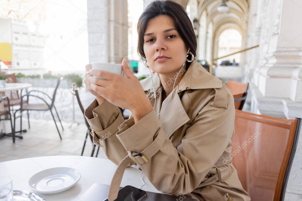 brunette woman in trench coat listening music and holding cup near smartphone and laptop in cafe terrace